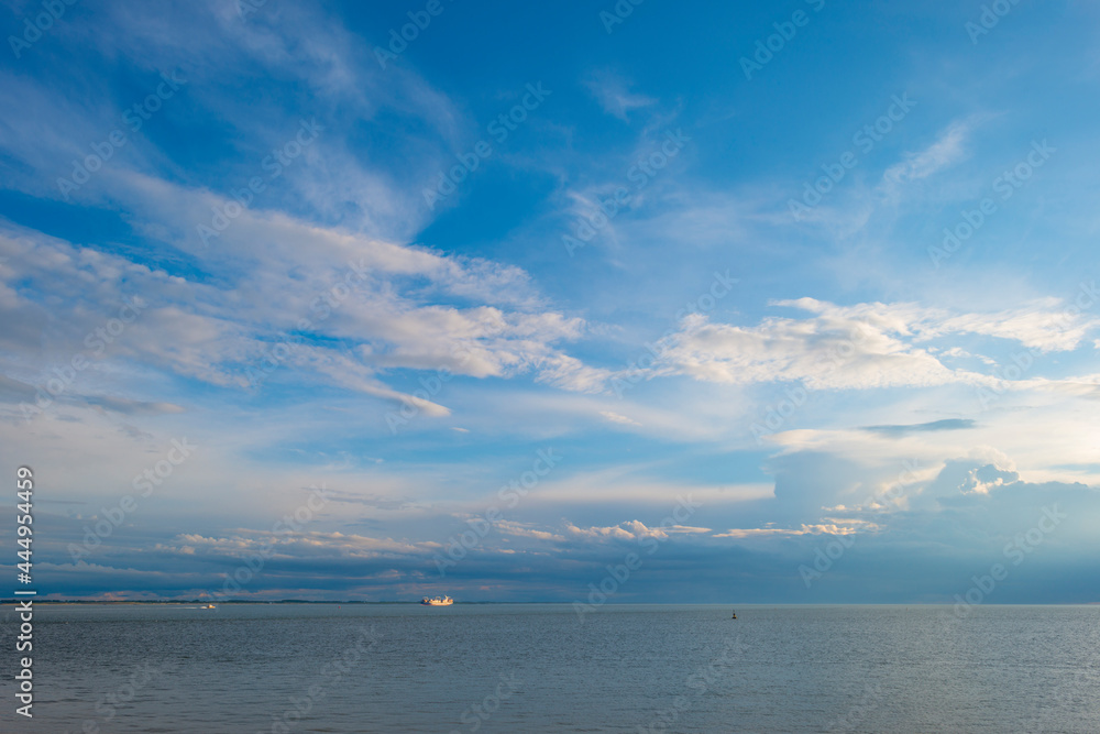 Obraz premium Sea along a coast under dark white clouds in a blue sky illuminated by the light of a colorful sundown sun in summer, Vlissingen, Walcheren, Zeeland, the Netherlands, July, 2021
