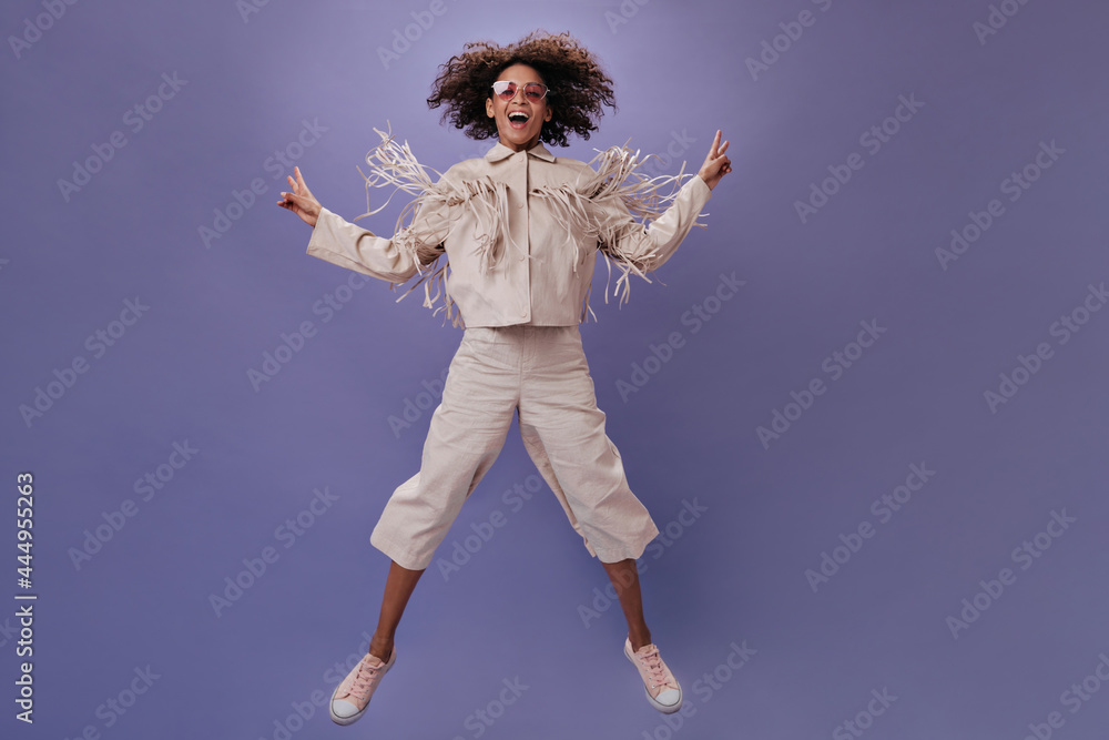Funny woman in jacket with fringe jumping on purple background. Smiling woman in beige pants and tee happily posing on isolated backdrop