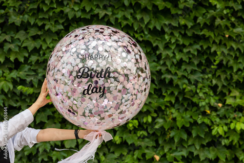 There's a wall of ivy. In front of it, a transparent balloon containing pink confetti is floating in the air and a happy birthday message is written.