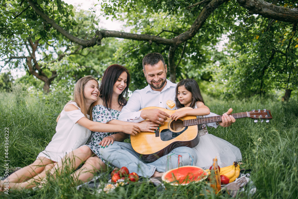 Happy family with cute daughters having picnic at green garden with guitar songs. Parents and two kids spending leisure time together with fun.