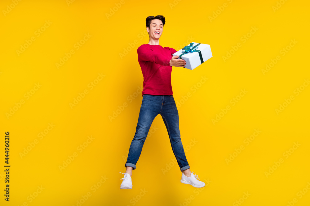 Photo of funny adorable young guy dressed red sweater holding present jumping high isolated yellow color background