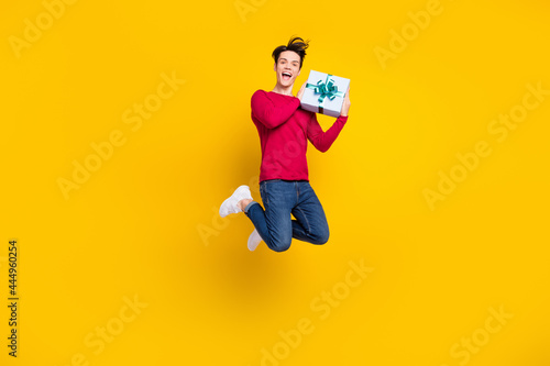Full length photo of funky excited young guy dressed red pullover holding present box jumping isolated yellow color background © deagreez