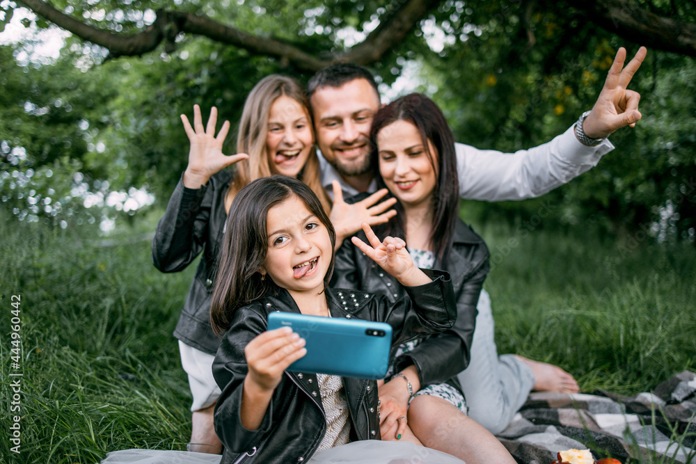 Fototapeta premium Cheerful parents and their cute daughters sitting together on green grass and using modern cell phone for taking self portrait. Concept of people, leisure and technology.