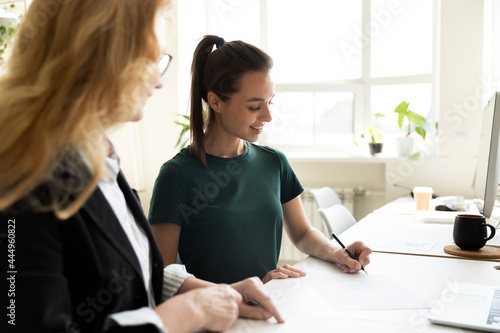 Mature business teacher supervising happy student writing notes. Middle aged mentor training intern at office workplace. HR manager helping new employee to fill and sign employment agreement photo