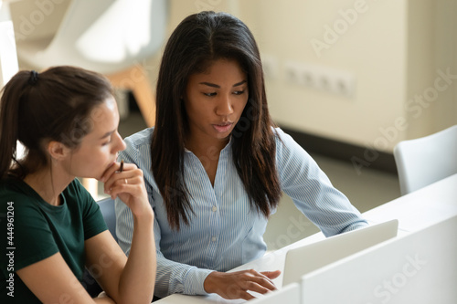 Serious diverse multiethnic student girls, interns collaborating on task at laptop together. Corporate mentor supervising new hired employee, helping with work task. Coworkers discussing project