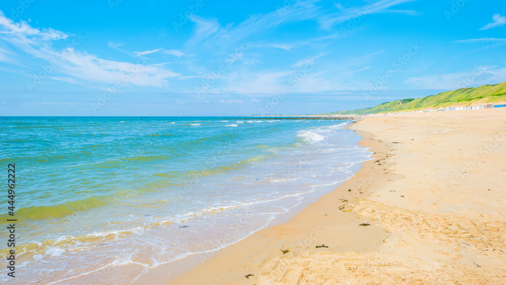 Obraz premium Sunlit waves on the yellow sand of a sunny beach along the North Sea illuminated by the light of a colorful sun and a blue cloudy sky in summer, Walcheren, Zeeland, the Netherlands, July, 2021 