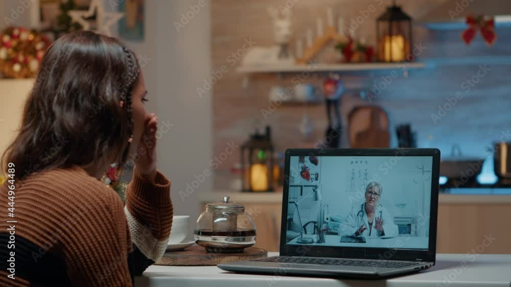 Woman with flu using telemedicine on laptop at home in christmas decorated kitchen. Sick young adult talking to doctor on internet for healing treatment against seasonal virus disease Woman with flu using telemedicine on laptop at home in christmas decorated kitchen. Sick young adult talking to doctor on internet for healing treatment against seasonal virus disease