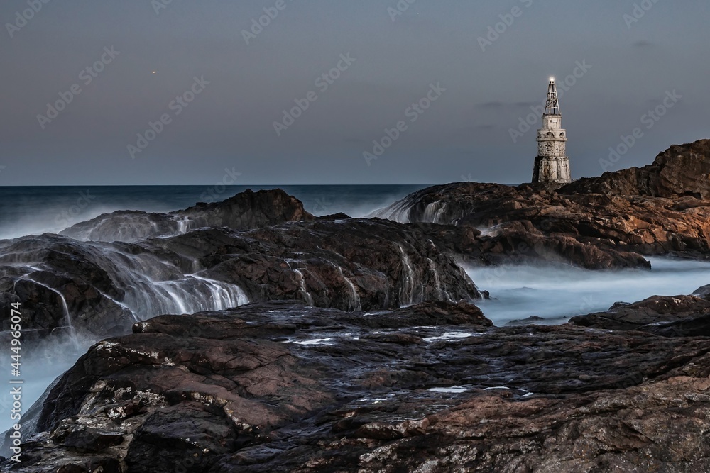 Attractive night seascape, sea waves splashing over rocks, shining lighthouse. Evening darkness, long exposure, blurry water trails and misty steam. White Southern Black Sea coast, Ahtopol, Bulgaria.