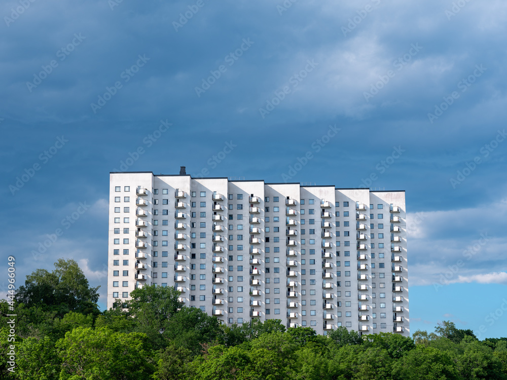 Whire apartment building with many balconies but no visible people. Green trees in foreground. Dark skies with thunder clouds. Sharp shadows on exterior from the sun.