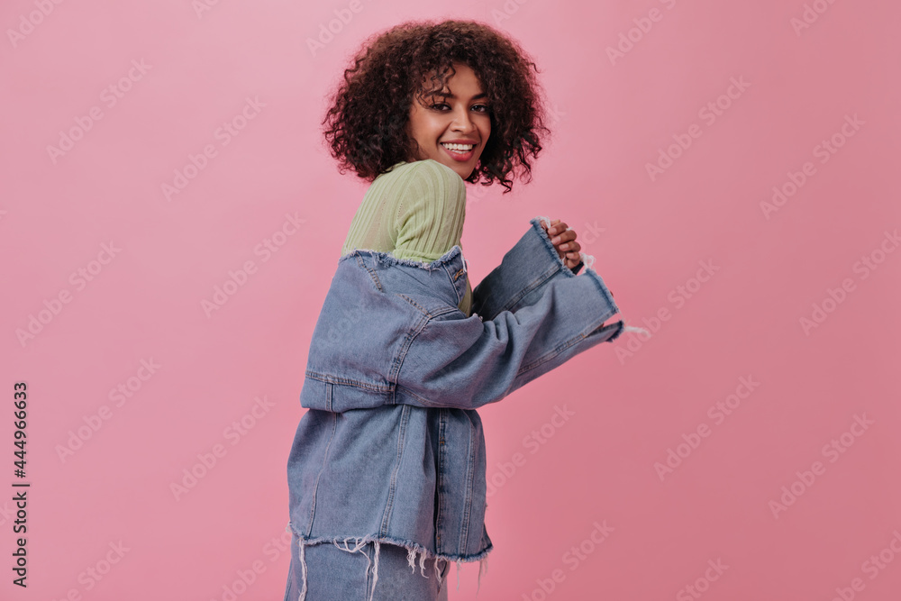 Charming woman in light denim outfit posing on pink background. Smiling girl in blue jacket and green tee laughing on isolated backdrop Charming woman in light denim outfit posing on pink background. Smiling girl in blue jacket and green tee laughing on isolated backdrop