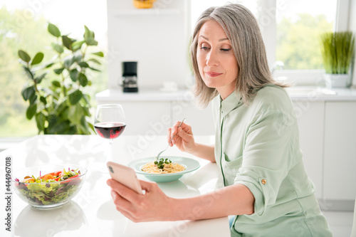 Portrait of attractive focused grey-haired woman eating fresh homemade dinner alone using device blogging at home light white indoors