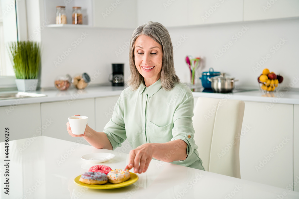 Portrait of attractive cheerful grey-haired woman drinking coffee eating bakery morning alone at home light white indoors Portrait of attractive cheerful grey-haired woman drinking coffee eating bakery morning alone at home light white indoors