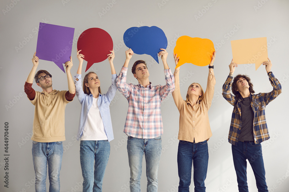 Group of young people looking up at multi colored empty cardboard and paper mockup speech bubbles they are holding standing on gray studio background. Sharing message and expressing opinion concept - obrazy, fototapety, plakaty 