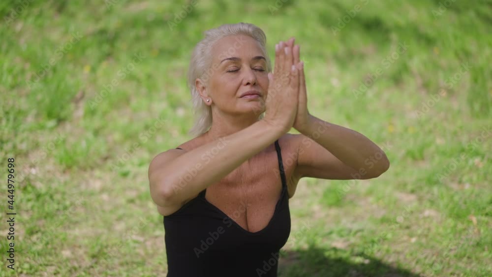 Medium shot of slim confident senior sportswoman raising hands in slow motion meditating on green summer lawn. Portrait of Caucasian yogi training in sunny park outdoors Medium shot of slim confident senior sportswoman raising hands in slow motion meditating on green summer lawn. Portrait of Caucasian yogi training in sunny park outdoors
