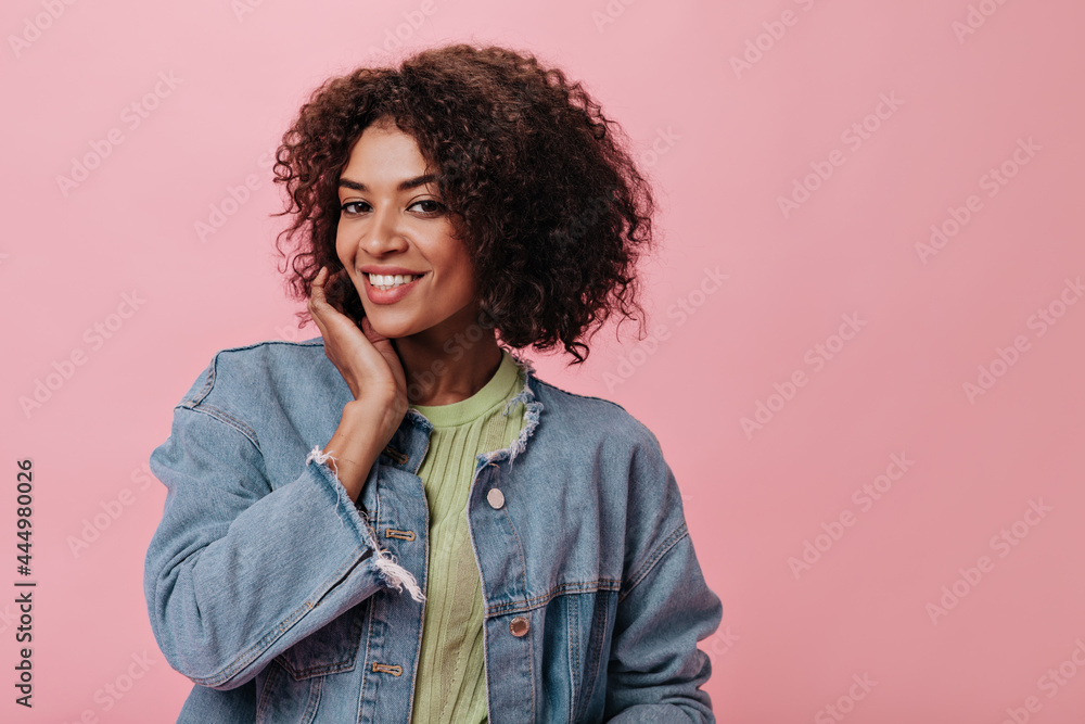 Portrait of curly woman in denim jacket on isolated background. Young pretty girl in green t-shirt smiling sincerely on pink backdrop Portrait of curly woman in denim jacket on isolated background. Young pretty girl in green t-shirt smiling sincerely on pink backdrop