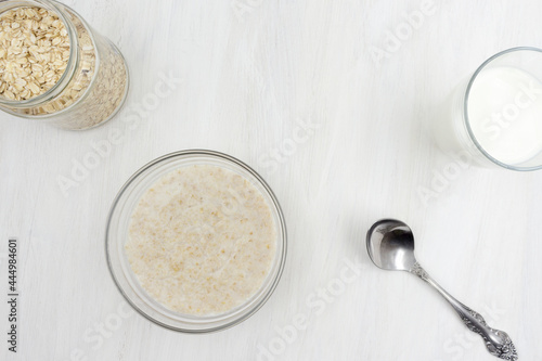 Oatmeal porridge cooked with milk, a glass of milk, a spoon, a plate on a white wooden table
