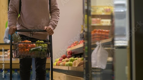 Vegetarian man carrying grocery cart full of fresh organic fruits and taking zucchini from store shelf. Young healthy eating shopper choosing vegetables and fruits in supermarket