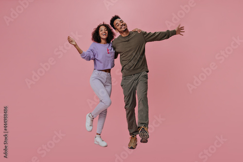 Happy couple in brown and purple outfits posing on pink background. Dark-skinned woman and brunette man smiling and hugging on isolated