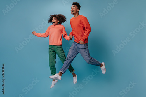 Surprised couple in orange sweatshirts jumping on isolated background. Shocked emotional man in red sweater and woman in green denim pants move on blue background