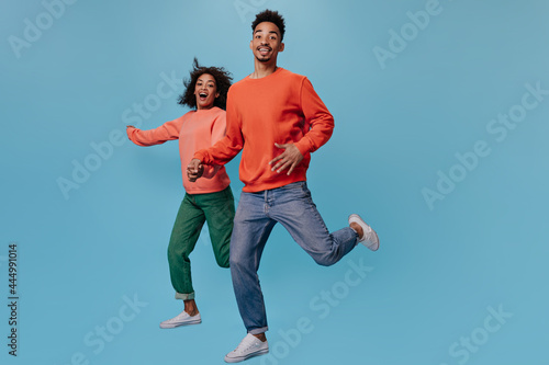 Cool man and woman in orange sweaters run on blue background. Emotional young girl and brunette man in jeans move and make steps