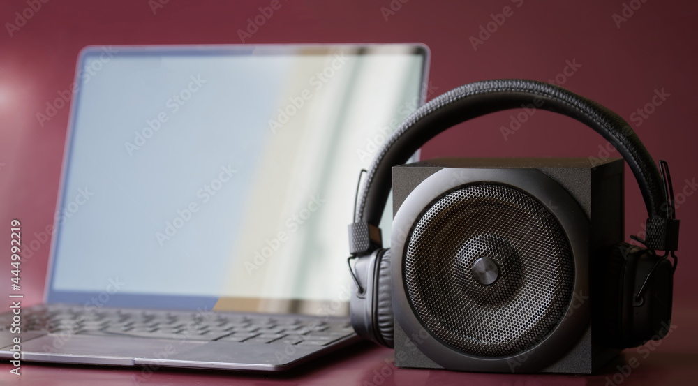 A square black audio speaker and wireless headphones next to a slim modern laptop. Concept for digital music and wireless audio gadgets. Burgundy background. Daylight A square black audio speaker and wireless headphones next to a slim modern laptop. Concept for digital music and wireless audio gadgets. Burgundy background. Daylight