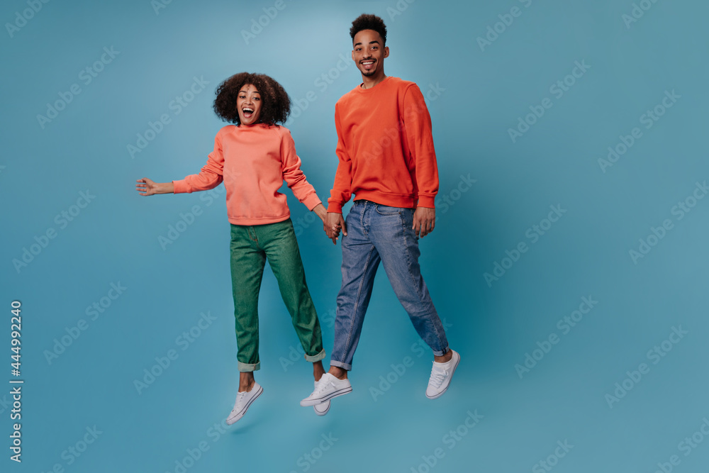 Funny man and woman in colorful outfits jumping on blue background. Portrait of young dark-skinned couple in movement on isolated backdrop