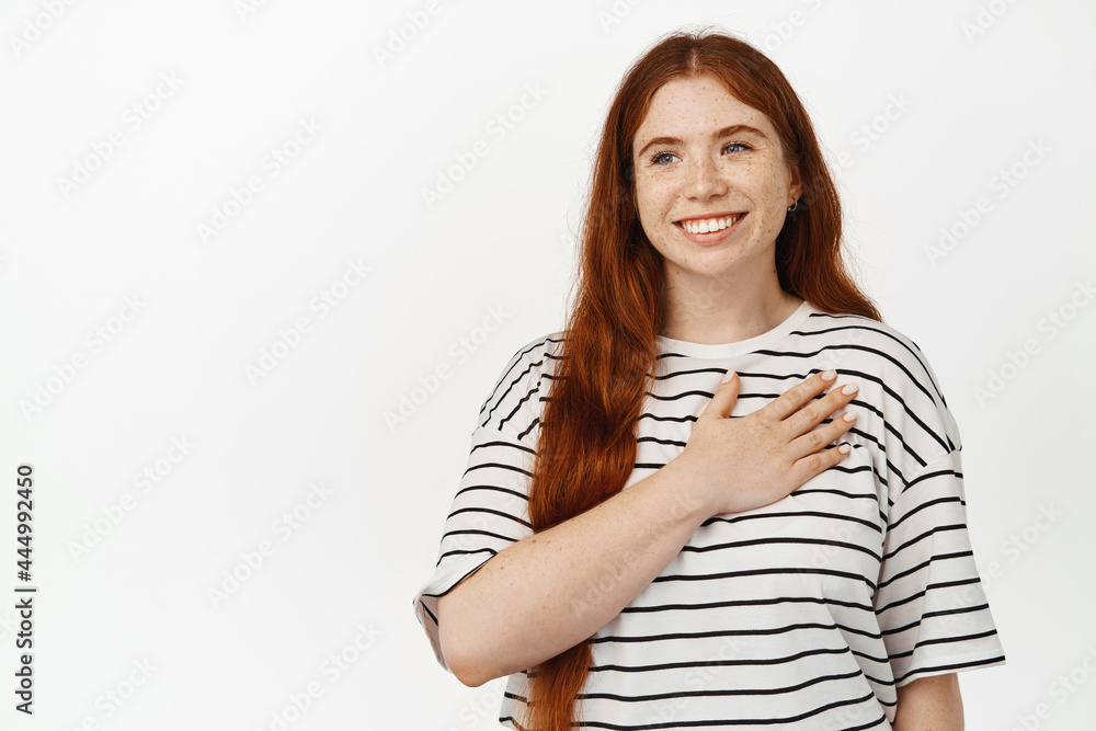Image of happy girl with ginger hair, hold hand on heart and smiling, looking away with pleased and grateful face expression, thanking for smth, standing against white background Image of happy girl with ginger hair, hold hand on heart and smiling, looking away with pleased and grateful face expression, thanking for smth, standing against white background