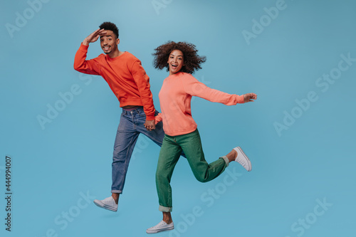 Dark skinned man and woman in orange sweatshirts running on blue background. Brunette girl in orange sweater and guy in jeans move on isolated