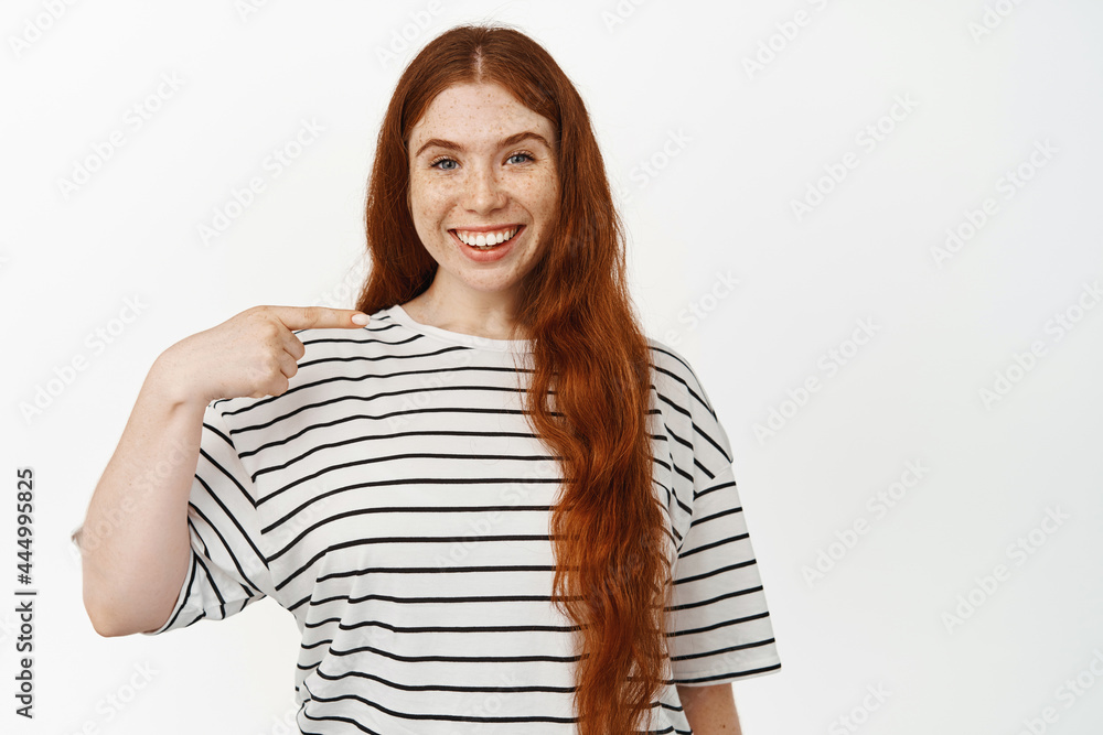 Image of smiling redhead girl with long natural hair, pointing at herself with pleased face expression, white teeth, showing something on her, standing against white background Image of smiling redhead girl with long natural hair, pointing at herself with pleased face expression, white teeth, showing something on her, standing against white background