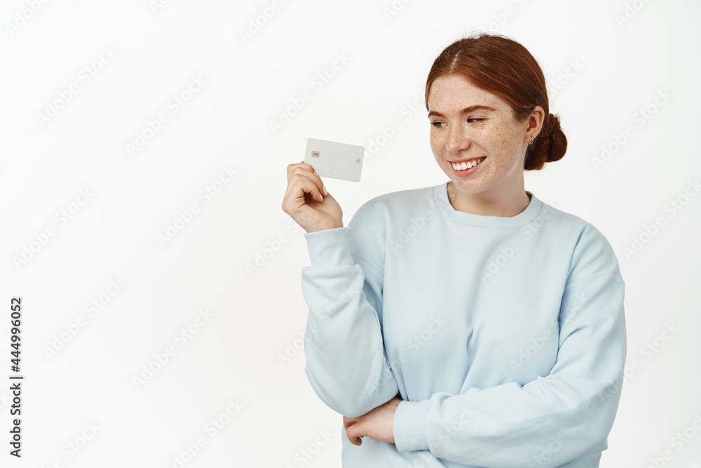 Beautiful young woman, girl with red hair holding credit discount card and smiling, looking pleased, paying in store contactless, standing carefree against white background Beautiful young woman, girl with red hair holding credit discount card and smiling, looking pleased, paying in store contactless, standing carefree against white background