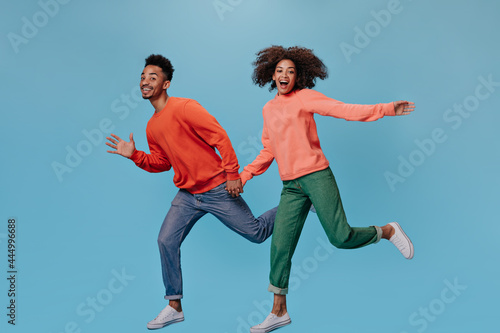 Happy guy and girl in stylish outfits moving on blue background. Dark-skinned positive woman in green denim pants and man in jeans run on isolated