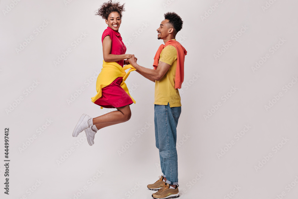 Man hold hands of his girlfriend while she jumping on white background. Brunette guy in jeans pose and active woman im red dress move up on isolated