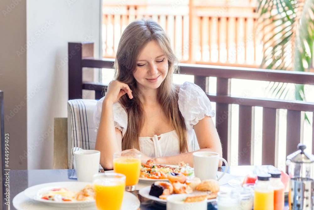 Young Smiling Woman Sitting on Breakfast with Plates Full of Food - Fresh Omelette, Vegetables, Fruits and Orange Juice in Glass. Morning Food in Luxury Hotel Young Smiling Woman Sitting on Breakfast with Plates Full of Food - Fresh Omelette, Vegetables, Fruits and Orange Juice in Glass. Morning Food in Luxury Hotel