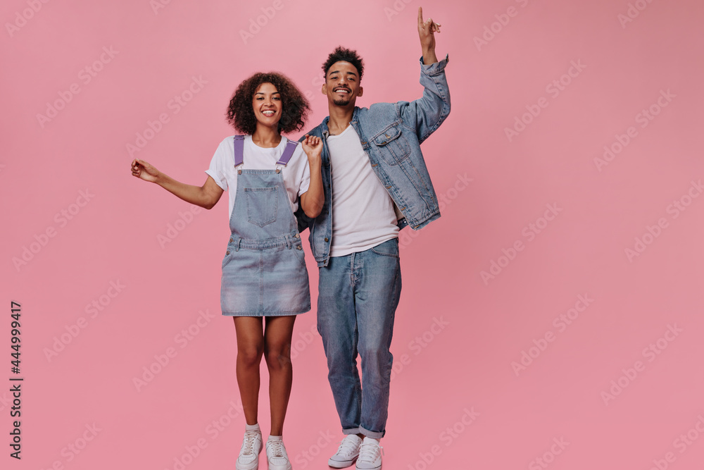 Obraz premium Brunette man and his girlfriend in jeans happily posing on pink background. Cool guy in denim jacket and girl in jumpsuit dance and smile