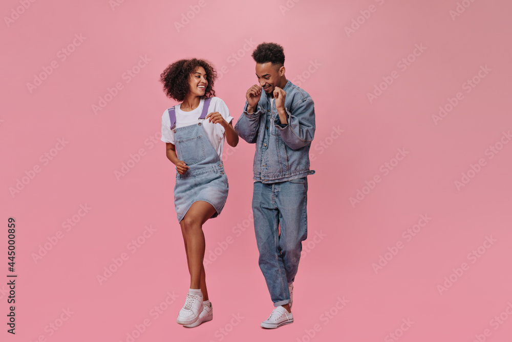Full length portrait of couple in denim outfits dancing on pink background. Joyful man in jeans and brunette lady in blue dress move and jump on isolated Full length portrait of couple in denim outfits dancing on pink background. Joyful man in jeans and brunette lady in blue dress move and jump on isolated