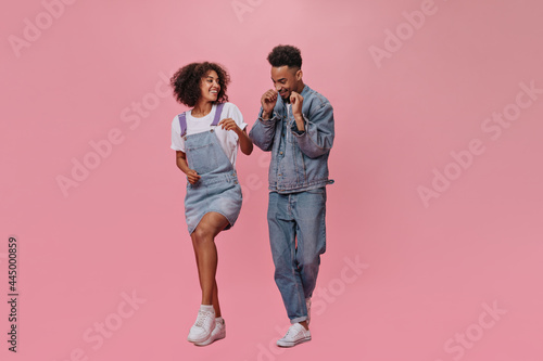 Full length portrait of couple in denim outfits dancing on pink background. Joyful man in jeans and brunette lady in blue dress move and jump on isolated