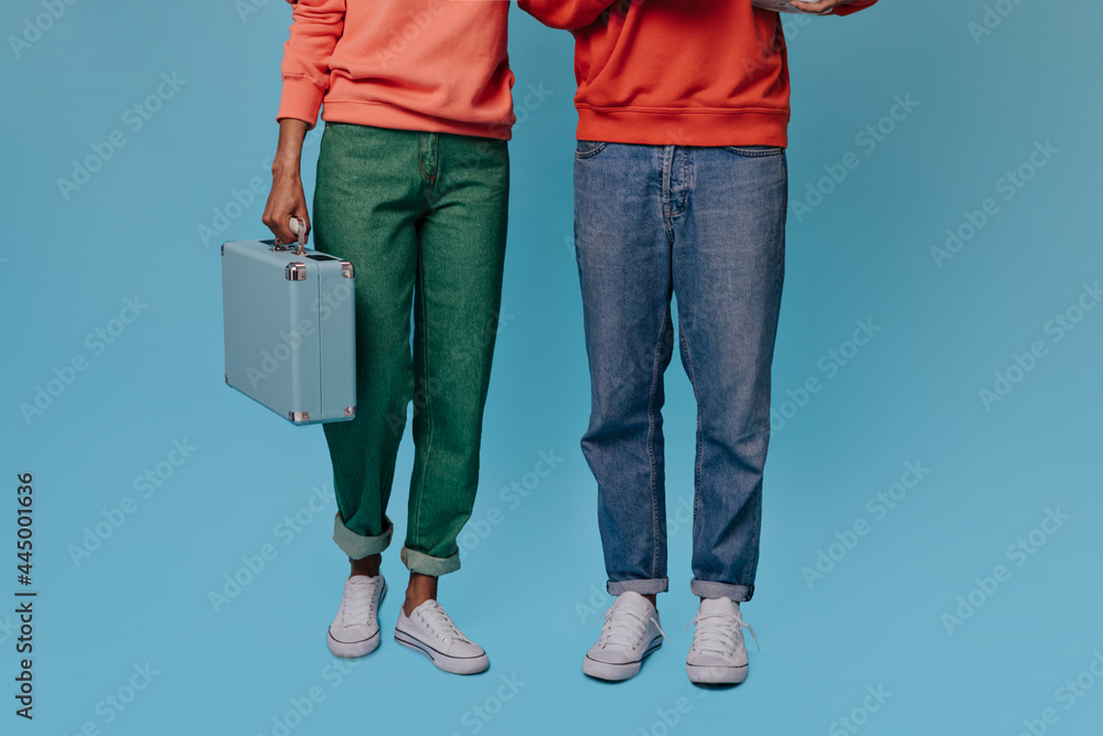 Naklejka premium Photo of legs of woman and man in jeans. Girl in baggy green denim pants holds blue suitcase on isolated background