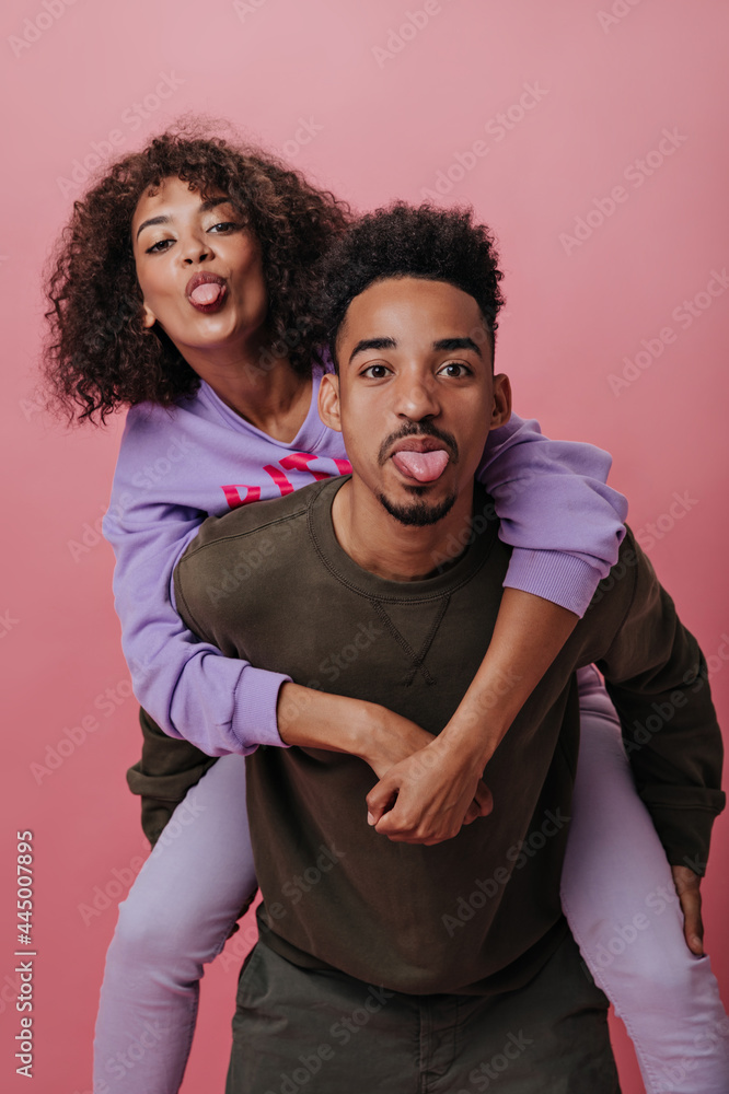 Obraz premium Brown-eyed man and woman having fun and showing tongues on pink background. Portrait of happy guy in brown outfit and girl hugging on isolated