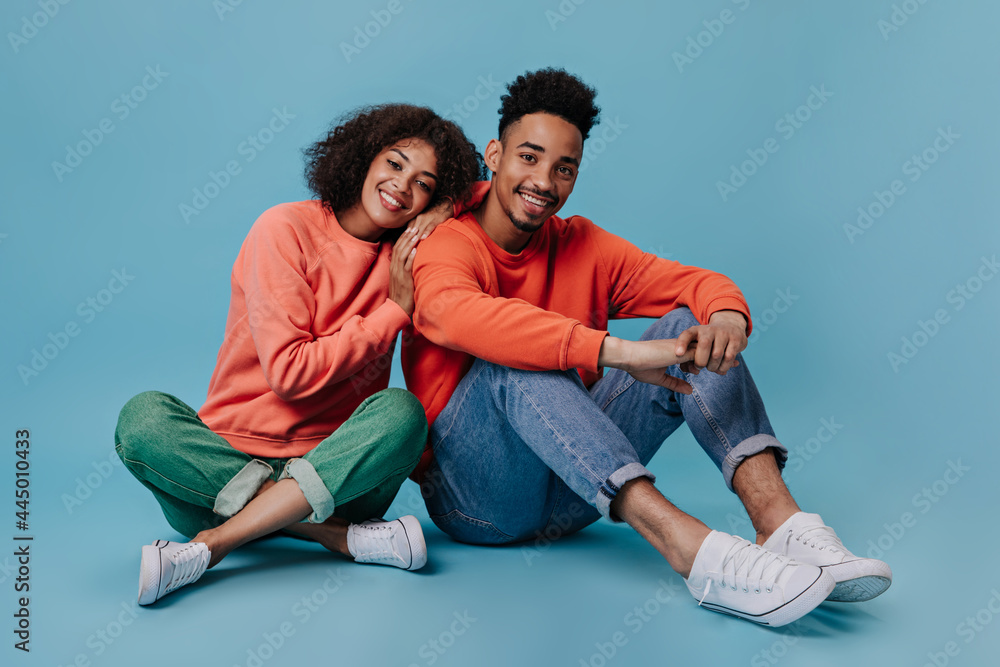 Young man and woman smiling and sitting on blue background. Portrait og guy in red sweatshirt and woman in gree jeans chilling on isolated Young man and woman smiling and sitting on blue background. Portrait og guy in red sweatshirt and woman in gree jeans chilling on isolated