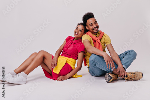 Man and woman in good mood sitting on white background. Cheerful girl in red dress and brunette guy in jeans smile on isolated