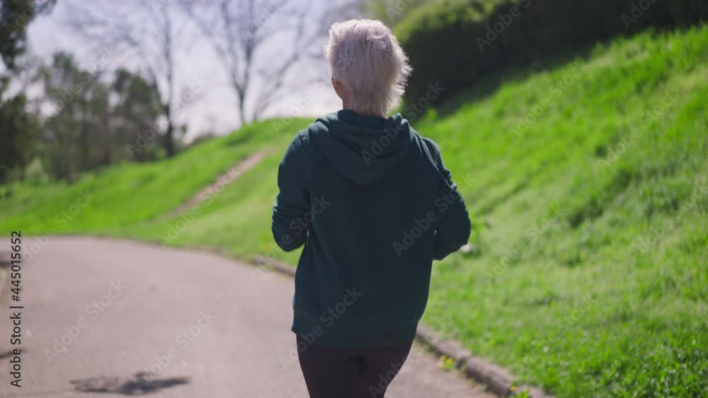 Live camera follows fit senior sportswoman jogging in slow motion in sunlight. Back view of slim Caucasian retiree running in spring summer park outdoors on sunny day. Sport and lifestyle concept Live camera follows fit senior sportswoman jogging in slow motion in sunlight. Back view of slim Caucasian retiree running in spring summer park outdoors on sunny day. Sport and lifestyle concept