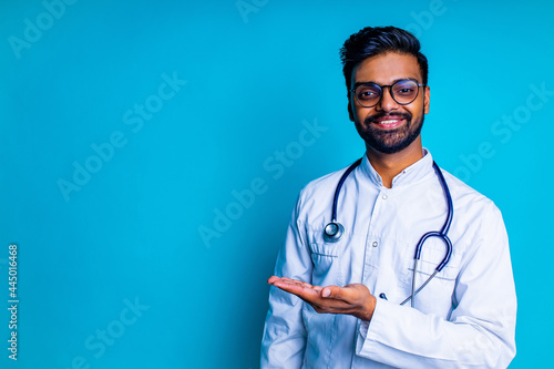 pakistanian doctor in white coat with stethoscope pointing at copyspace with hand mock up over blue studio background photo