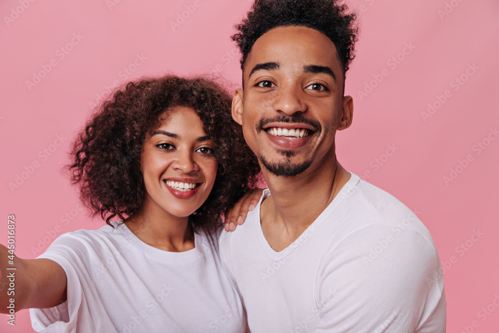 Charming woman and man dressed in white T-shirts smiling and taking selfie. Portrait of cheerful curly girl and dark-skinned girl in tee posing on pink background Charming woman and man dressed in white T-shirts smiling and taking selfie. Portrait of cheerful curly girl and dark-skinned girl in tee posing on pink background
