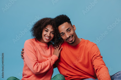Brown eyed guy and his curly lady hugging and smiling on blue background. Charming woman in orange sweater and her boyfriend pose on isolated