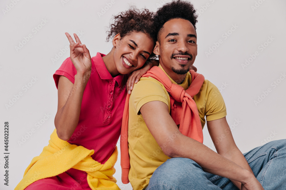 Naklejka premium Woman in red dress shows peace sign, winks and hugs her boyfriend. Happy lady in shirt and young man in jeans smile on white background