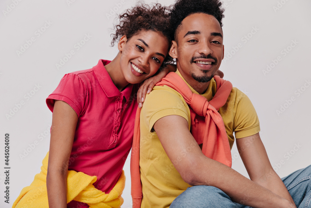 Naklejka premium Dark-skinned guy and girl sitting on white background. Joyful woman in red shirt and brunette man in yellow tee smile on isolated