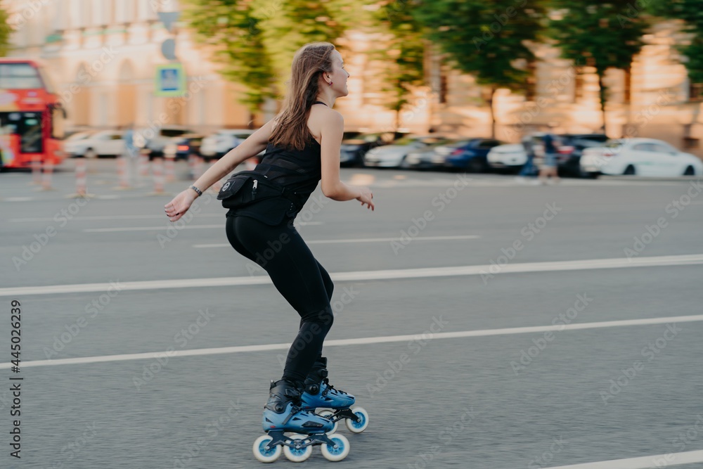 Active lifestyle and hobby concept. Sporty young woman does sport outdoors rides rollerblading dressed in sportsclothes enjoys fitness activities poses in urban place on road. Rollerskating.