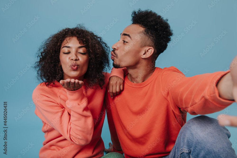 Girl in orange sweatshirt blows kiss while her boyfriend taking selfie. Happy young man in jeans and curly woman sit and pose on blue background Girl in orange sweatshirt blows kiss while her boyfriend taking selfie. Happy young man in jeans and curly woman sit and pose on blue background