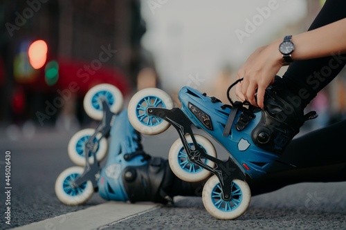Cropped shot of unrecognizable woman puts on rollerblades poses outdoors on asphalt against blurred background. Unknown person wears rollerskates spends free time actively tries new purchase