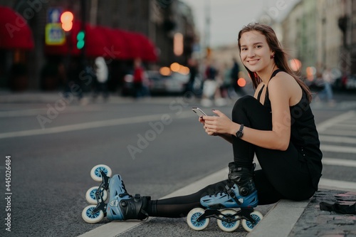People outdoor activities and recreation concept. Horizontal shot of active slim woman being in good physical shape rides rollerblades uses smartphone sends text messages online poses outside © VK Studio